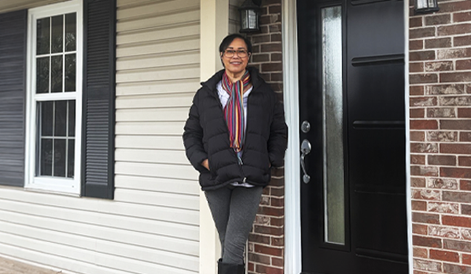 A woman smiles in front of a black door wearing a dark jacket and scarf a window with black shutters is to her left