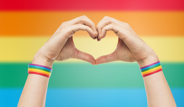 Hands make a heart sign in front of a rainbow background and there are rainbow wrist bands